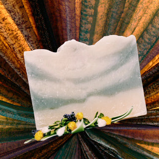 A raw bar of Simply Hypnotic Organic Soap with Geranium, Cedarwood & Patchouli Essential Oils, displayed on a coloful background with small flowers